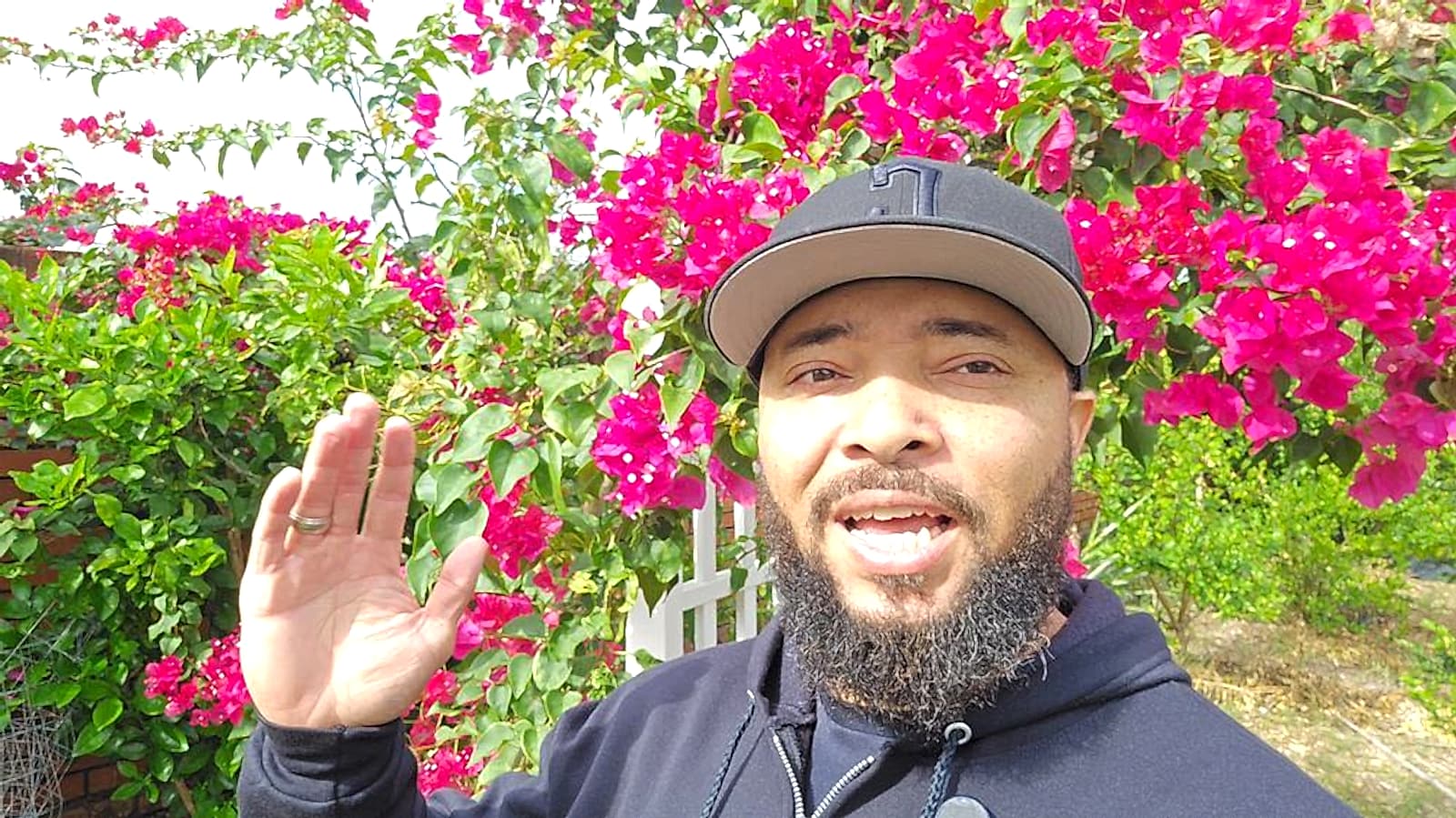 Jermaine waving to the camera in front of bright pink bougainvillea in his yard